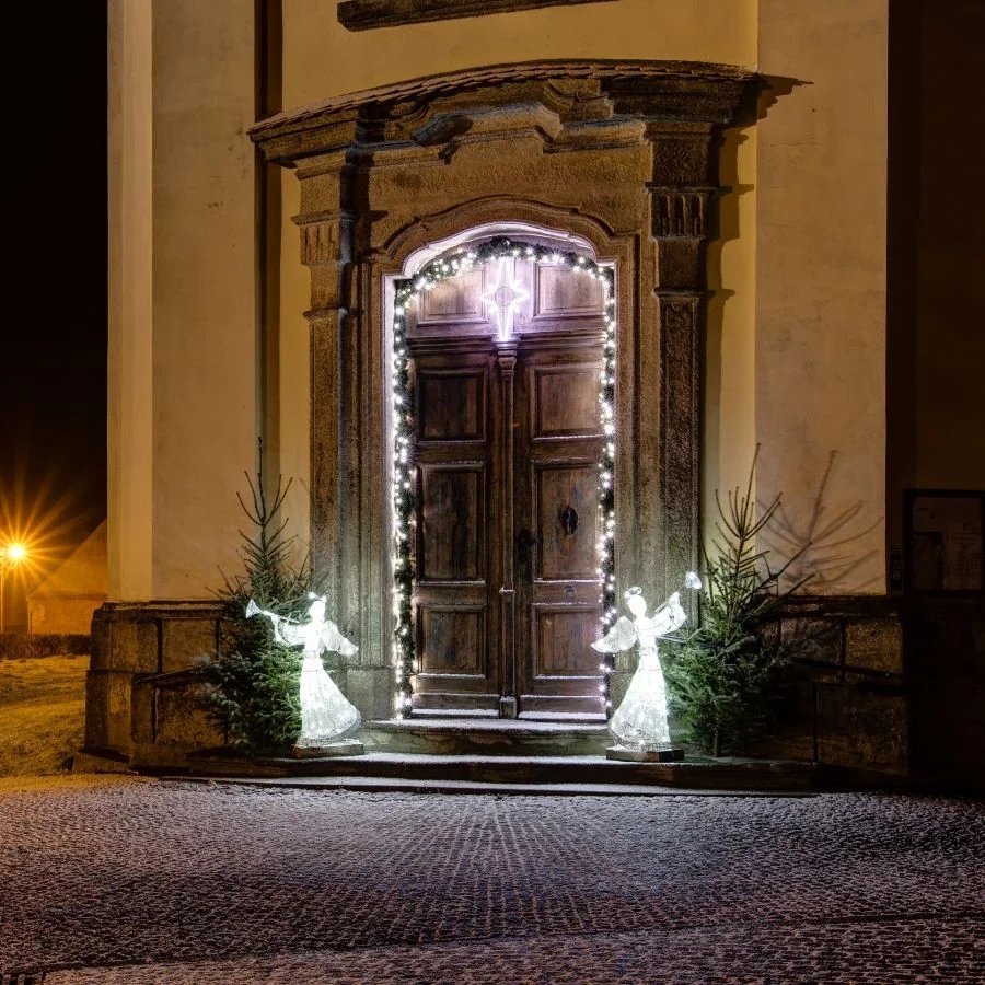 Two ice white LED angels with trumpets by a baroque door with Christmas trees, a night shot of the entrance